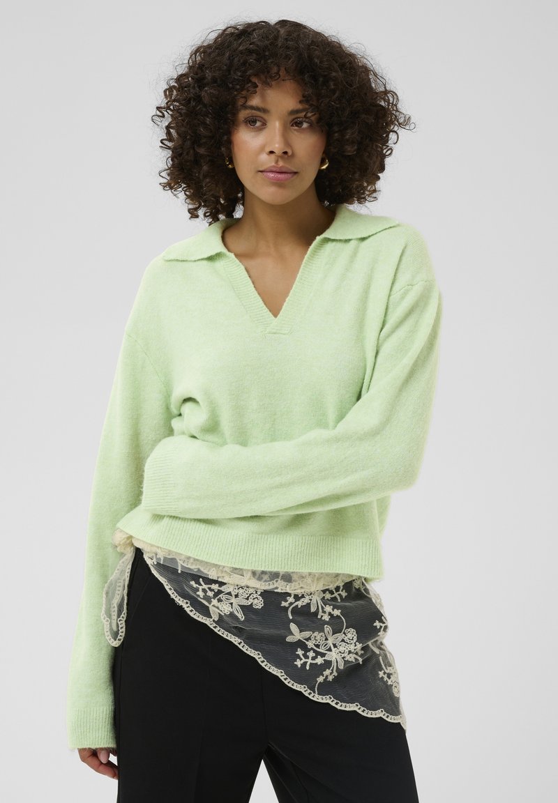 Pull en maille vert clair avec col en V et poignets côtelés, superposé à un détail en dentelle noire transparente avec des motifs floraux.