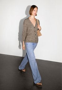 Brązowy sweter z dzianiny z ciemnymi guzikami, połączony z jasnoniebieskimi dzwonami jeansami i brązowymi sandałami. Modelka nosi małą beżową torebkę.