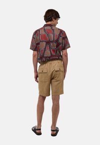 Chemise à manches courtes, à motifs brun foncé et beige avec des accents blancs, associée à un short beige comportant deux poches arrière, et des sandales noires.