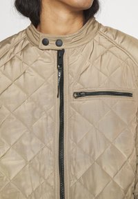 Beige quiltad jacka med en glänsande yta, som har en svart dragkedja framtill, tryckknappar vid kragen och en dragkedjeförsedd bröstficka.