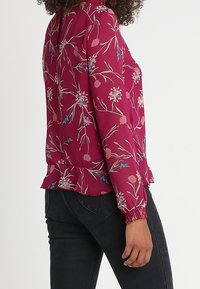 Vinröd blus med blommönster, långärmad, med rynkade ärmslut och volangkant. Har rosa, blå och gröna blommönster på ett texturerat tyg.