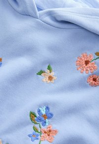 Tissu bleu clair avec de petites fleurs brodées en rose, bleu, beige et feuilles vertes, montrant la texture et les détails cousus.