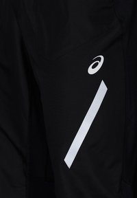 Gros plan d’un pantalon de sport noir avec le logo blanc Asics et une bande diagonale réfléchissante sur la jambe.