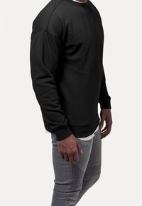 Felpa nera con scollo rotondo e polsini a costine, realizzata in un tessuto liscio e strutturato. Indossata sopra una camicia bianca e abbinata a jeans grigi.