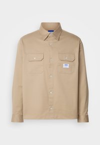 Chemise beige à manches longues boutonnée avec deux poches à rabat à l’avant, dont une poche avec une petite étiquette blanche avec le texte bleu « HUGO ».