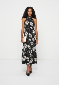 Vestido maxi preto com decote halter, apresentando um padrão floral branco, tecido fluido e uma silhueta elegante. Combinado com uma pequena carteira bege e saltos pretos.