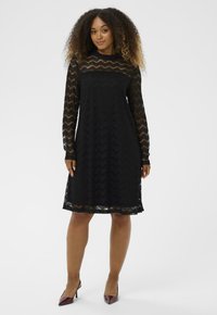 Crna čipkasta haljina dugih rukava, s valovitim uzorkom i labavim krojem, do sredine butina. Uparena s bordo potpeticama.