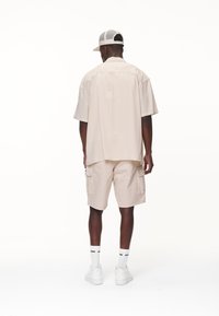 Beige kortærmet skjorte parret med matchende cargo shorts, der har lommer. Hvide sneakers og en hvid mesh-cap fuldender outfittet.