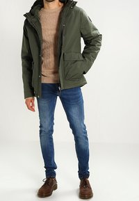 Olivgrön isolerad jacka med dragkedja framtill, två sidfickor och huva. Bärs över en beige stickad tröja och blå jeans. Bruna skor.