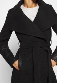 Abrigo negro con un cuello estructurado, cinturón en la cintura y mangas de cuero acanaladas. Presenta una tela texturizada y bolsillos laterales.
