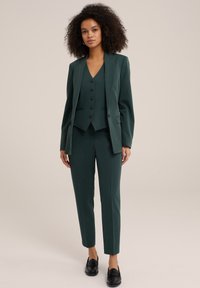 Tmavě zelený oblek na míru s přiléhavým sako s klopovým límcem, odpovídající vestou s knoflíky a slim-fit kalhotami. Dokončeno černými mokasínami.