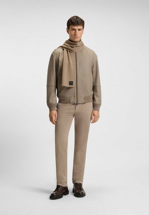 Veste bomber en cuir tanné, pantalon ajusté assorti et écharpe beige. Des bottes à lacets marron complètent le look, avec une étiquette de marque discrète sur l'écharpe.