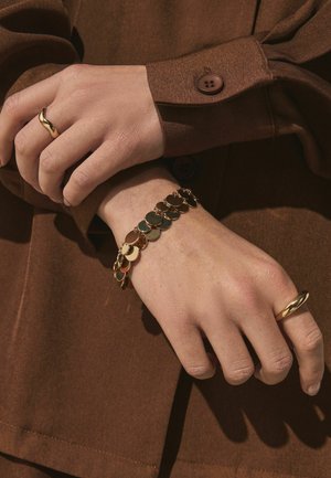 Hands crossed wearing gold rings and a gold bracelet with round discs, against a brown long-sleeve shirt with a buttoned cuff.