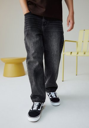 Persona che indossa scarpe da ginnastica nere e jeans neri larghi, in piedi vicino a una sedia gialla e uno sgabello rotondo giallo su un pavimento bianco.