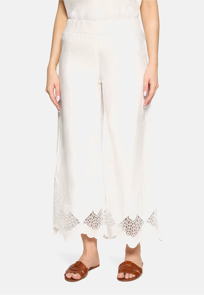 Femme portant un pantalon blanc à jambes larges avec une bordure en dentelle à l'ourlet et des sandales plates marron, debout devant un fond uni.
