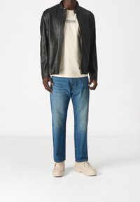 Hombre con chaqueta de cuero negra, camiseta gráfica beige, jeans azules y botas de cordones beige claro de pie contra un fondo blanco.