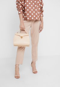 Kvinna i beige byxor och en rosa blus med prickar, som håller en beige handväska och har på sig beige högklackade sandaler mot en vit bakgrund.