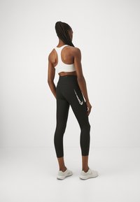 Svarta leggings med hög midja och en vit Nike-logotyp på sidan, tillsammans med en vit sport-bh, sett bakifrån.