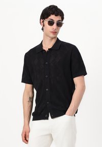 Camisa negra de manga corta con botones y un patrón de diamantes, que presenta un cuello y un diseño de punto texturizado, combinada con pantalones blancos.