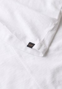 Camiseta blanca de algodón con una textura que parece ligeramente arrugada, con una pequeña etiqueta negra en un lado con texto en naranja.