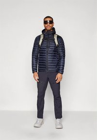 Marinblå vadderad jacka med dragkedja fram, lättviktigt material och avtagbar huva; kombinerad med mörka byxor och grå sneakers. Svarta solglasögon och en ryggsäck.