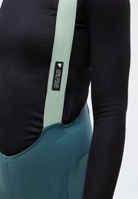 Κοντινό πλάνο ενός ατόμου που φοράει ποδηλατικά σορτς bib σε τιρκουάζ χρώμα με λουριά ανοιχτού πράσινου πάνω από μια μαύρη υφασμάτινη μακρυμάνικη βάση.