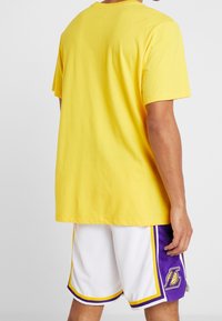Żółta bawełniana koszulka z krótkim rękawem, zestawiona z białymi siateczkowymi szortami z fioletowo-żółtymi akcentami i logo na dolnej stronie.