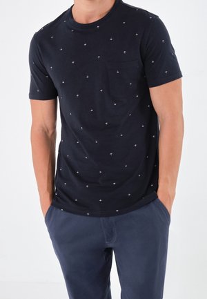 Camiseta de algodón azul marino con pequeños patrones de palmeras blancas, cuello redondo, mangas cortas y un bolsillo en el pecho izquierdo, combinada con pantalones azul marino.