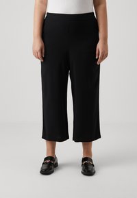 Pantalons noirs courts avec une texture lisse, ceinture élastique et coupe ample, associés à des mocassins noirs brillants ornés d'une chaîne dorée.