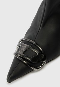 Fekete, hegyes orrú cipő sima bőrből, texturált ezüst csattal a lábfejen, oldalán varrott részletekkel.