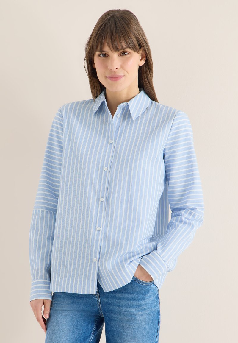 Femme aux cheveux bruns portant une chemise à rayures verticales bleu clair et blanc et un jean bleu, debout avec une main dans la poche contre un mur uni.