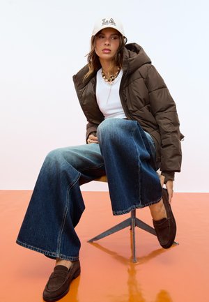 Giacca a vento marrone, t-shirt bianca, jeans a gamba larga blu, mocassini marroni e cappellino beige con testo "LA". Seduto su una sedia di metallo davanti a uno sfondo arancione.
