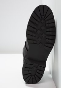 Botte noire avec une semelle épaisse en caoutchouc rainuré affichant un motif de semelle en forme de croix, posée debout contre un mur et une surface blancs.