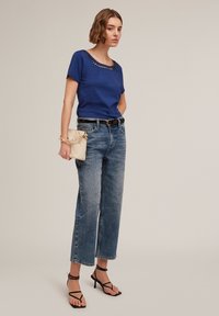 Camiseta azul de manga corta con escote recortado, combinada con vaqueros de pierna ancha de color azul claro y tacones negros con tiras. Sosteniendo un bolso beige texturizado.