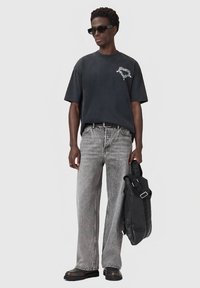 Άνδρας φορεμένος με μαύρα γυαλιά ηλίου, υπερμεγέθη σκουρόχρωμη μπλούζα, γκρίζα τζιν, μαύρα παπούτσια, κρατώντας μαύρη τσάντα απέναντι από ουδέτερο ανοιχτό φόντο.