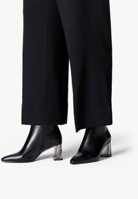 Stivali neri alla caviglia in pelle liscia, con punta appuntita e tacco a blocco trasparente. Indossati con pantaloni larghi neri, che mettono in evidenza gli stivali.