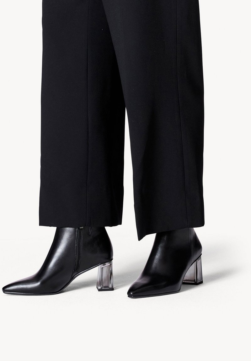 Stivali neri alla caviglia in pelle liscia, con punta appuntita e tacco a blocco trasparente. Indossati con pantaloni larghi neri, che mettono in evidenza gli stivali.