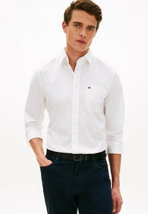 Młody mężczyzna w białej koszuli z guzikami i małym logo na kieszeni, granatowych spodniach oraz czarnym pasku, stojący z jedną ręką w kieszeni.