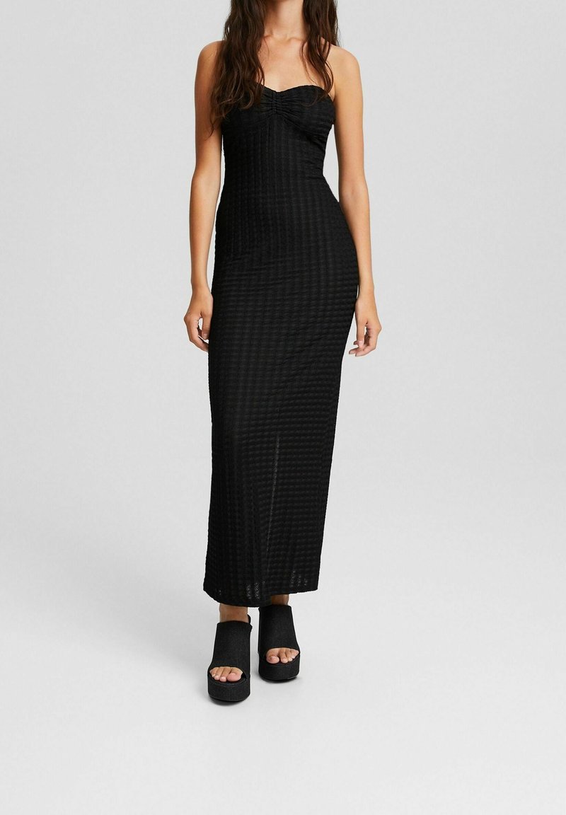 Femme portant une robe longue noire bustier avec une texture subtile et des chaussures à plateforme noires ouvertes sur les orteils, debout devant un fond uni.