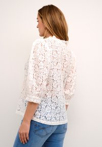 Blouse en dentelle blanche à manches trois-quarts, avec un col volanté et un motif floral. Associée à un jean en denim bleu.