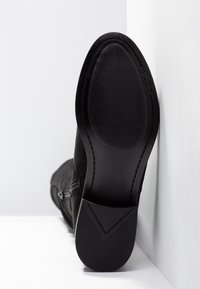 Semelle d'une bottine noire à cheville avec une semelle texturée et une fermeture éclair sur le côté, placée debout contre un coin blanc.