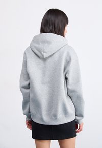 Donna con capelli scuri di media lunghezza, indossa una felpa grigia con cappuccio e una gonna nera, vista di spalle su uno sfondo chiaro e semplice.