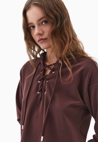 Brązowy hoodie z dekoltem na sznurki, wyposażony w srebrne akcenty metalowe. Miękka tkanina, luźny krój i gładka textura.