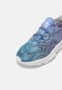 Zapatilla con un upper textil en degradado azul y púrpura que presenta patrones de olas, media suela blanca y cordones púrpuras. Logo de Adidas en la lengüeta.