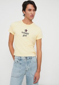 Joven vistiendo una camiseta amarilla pálido de "tommy girl" y jeans de tiro alto de color azul claro, de pie con una mano en el bolsillo, mirando hacia un lado.