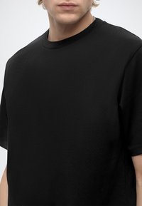 Young person wearing a plain black short-sleeve crew neck t-shirt against a plain light background.