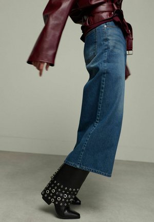 Wijd gesneden blauwe denim jeans gecombineerd met zwarte leren enkellaarzen met zilveren oogje accenten. De outfit omvat een bordeaux leren top.