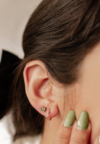 Pendiente de aro dorado con acentos multicolores al lado de un pequeñostud de piedra preciosa verde, usado en una oreja lisa. Uñas verde oliva tocan la piel.