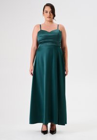 Langes satin Kleid in dunklem Petrol mit einem taillierten Oberteil, verstellbaren Spaghettiträgern und einem schwingenden Rock. Getragen mit schwarzen spitzen Absätzen.