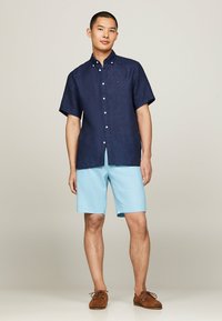 Camicia a maniche corte button-up blu marino abbinata a pantaloni corti azzurri. Scarpe marroni stringate completano il look estivo. Tessuto morbido con una vestibilità casual.